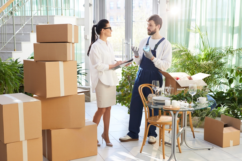 female event manager with pony tail making notes in paper while holding clipboard and discussing goods replacement with mover while preparing space for event.