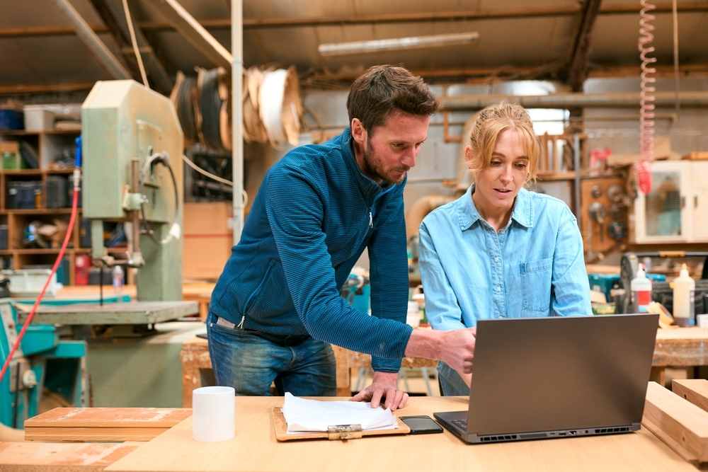 Male And Female Carpenters Working In Woodwork Workshop Using Laptop Together