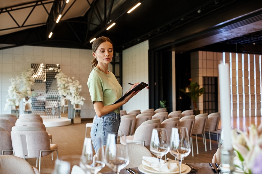 young event manager with clipboard looking at table with festive setting in modern event hall